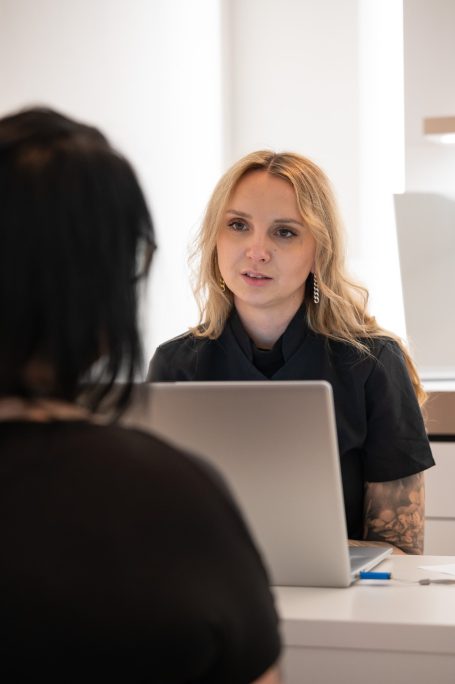 Blonde Frau am Computer, spricht mit einer Person im Vordergrund.