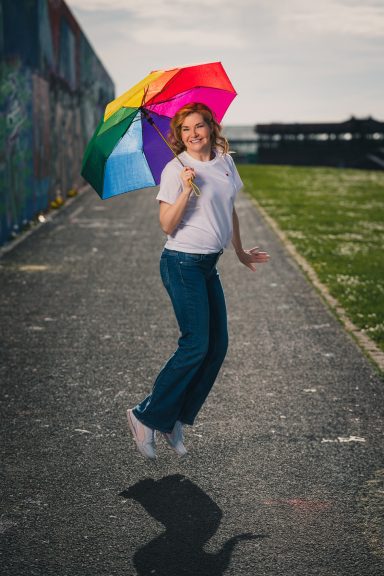 Frau mit buntem Regenschirm springt fröhlich auf einem Weg.
