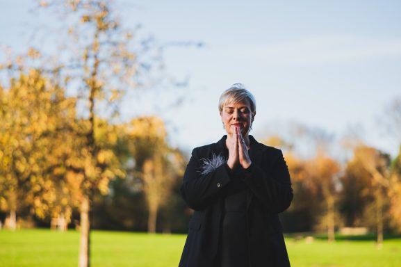 Eine Person steht im Freien mit gefalteten Händen, umgeben von herbstlichen Bäumen.