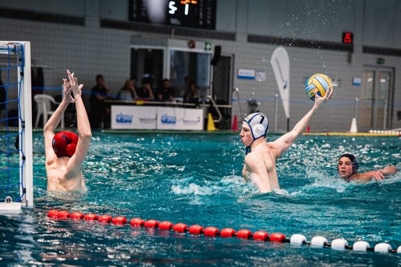 Wasserballspieler wirft Ball auf das Tor, während Gegner versucht, zu verteidigen.