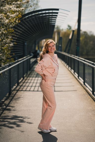 Frau in einem rosafarbenen Anzug steht auf einer Brücke mit Bäumen im Hintergrund.