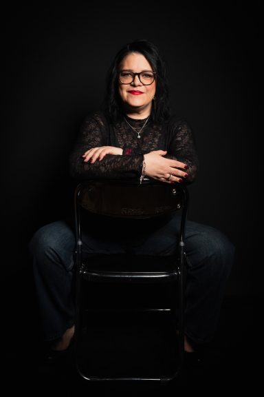 Frau mit Brille in schwarzem Oberteil sitzt lächelnd auf einem Stuhl vor schwarzem Hintergrund.