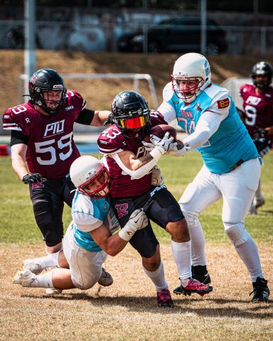 Zwei Spieler im Football kämpfen um den Ball, während ein weiterer Spieler tackelt.