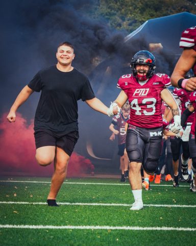 Ein Spieler und ein Fan laufen gemeinsam auf das Spielfeld, umgeben von Rauch.