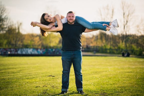 Mann hebt lächelnde Frau auf seinen Schultern in einer sonnigen Freifläche.