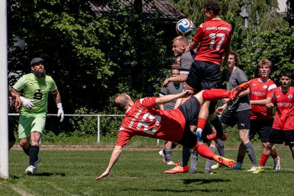 Fußballspieler springt, um den Ball im Torraum zu erreichen; Torwart bereit für den Schuss.