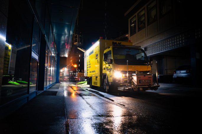 Gelbes Lieferfahrzeug auf einer nassen Straße bei Nacht, mit beleuchteten Fassaden im Hintergrund.