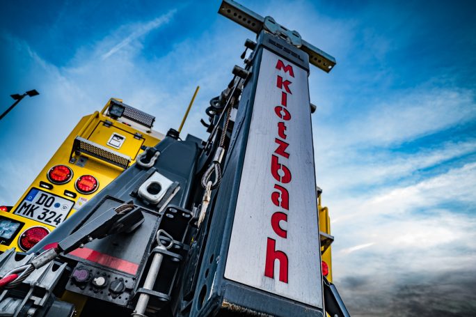 Gelbe Maschine mit hoch aufragendem Schild "Mikrolzbach" vor einem blauen Himmel.