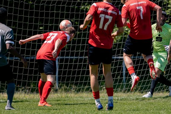 Spieler in roten Trikots springen, während ein Kopfball auf das Tor zugeht.