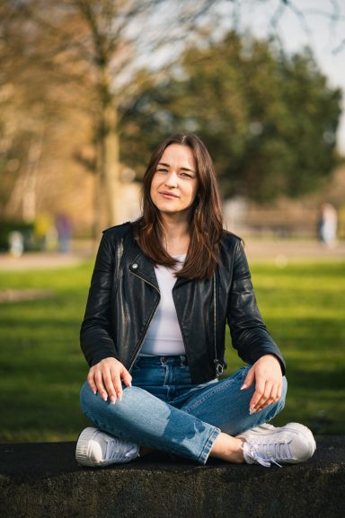 Frau mit Brille und Lederjacke sitzt entspannt im Freien auf dem Boden.