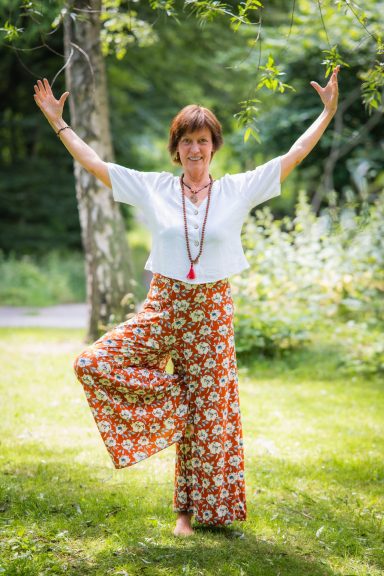 Frau in weiter Hose und Bluse, steht mit erhobenen Armen und positiver Ausstrahlung im Freien.
