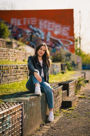 Junge Frau in Lederjacke sitzt auf einer Treppe vor einer bunten Wandmalerei.