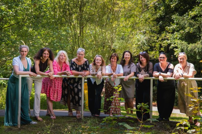 Eine Gruppe von neun Frauen lächelt am Geländer in einem grünen Garten.