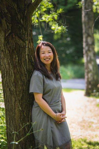 Frau lächelt, lehnt an einem Baum in einer grünen, sonnigen Umgebung.