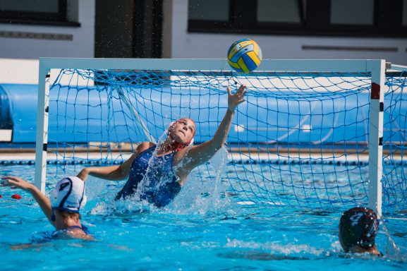 Wasserballspiel: Spielerin springt, um den Ball über das Tor zu schlagen.