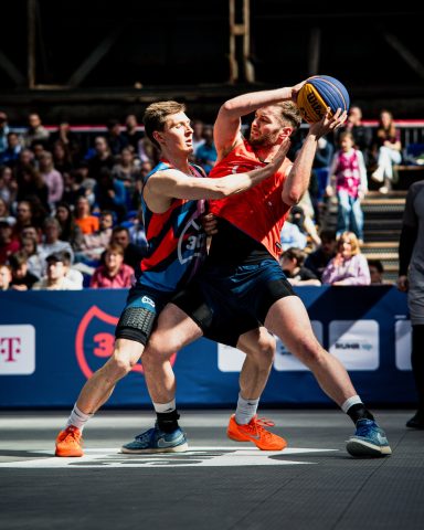 Zwei Basketballspieler kämpfen um den Ball in einem spannenden Spiel. Zuschauer im Hintergrund.