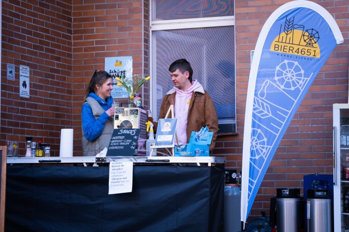 Zwei Personen stehen an einem Tisch, besprechen Informationen, im Hintergrund ein Banner.