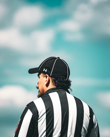 Ein Schiedsrichter mit gestreiftem Shirt und schwarzer Kapuzenmütze vor einem klaren Himmel.