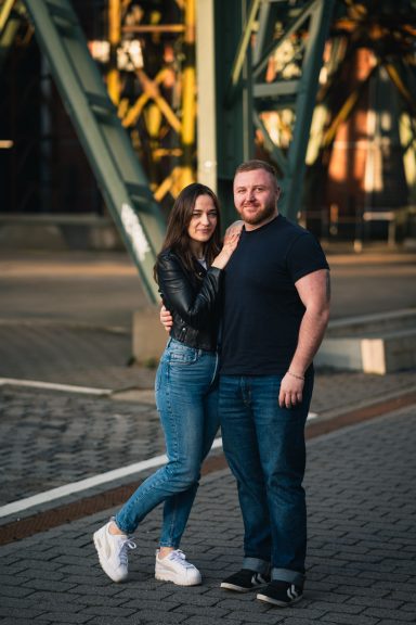 Paar steht lächelnd in urbaner Umgebung, umarmt sich vor einer industriellen Kulisse.