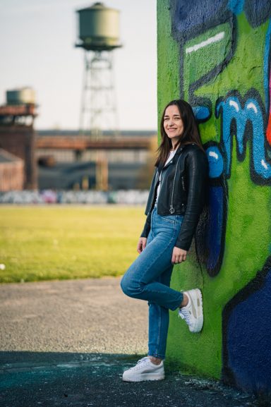 Junge Frau in Lederjacke und Jeans lehnt an einer bunten Wand in einem Industriegebiet.