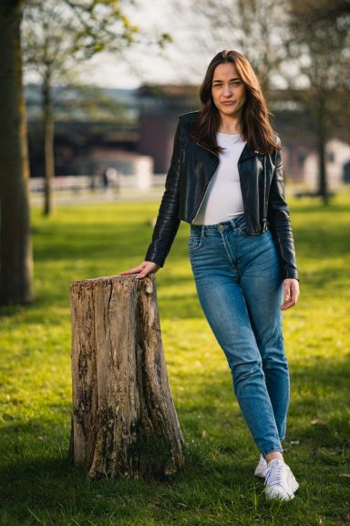 Junge Frau in einer Lederjacke lehnt an einem Baumstumpf im Freien.