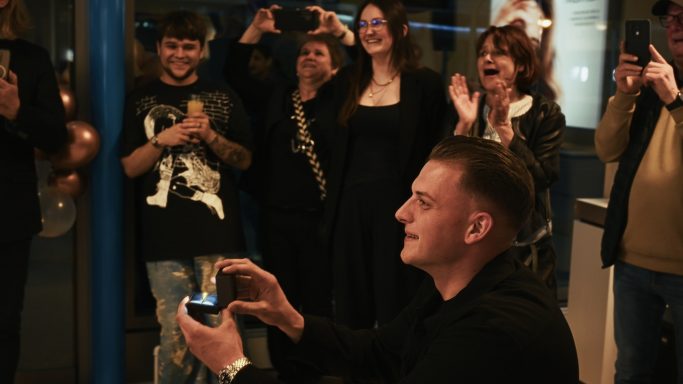 Ein Mann kniet und hält einen Ring, während Umstehende begeistert reagieren.