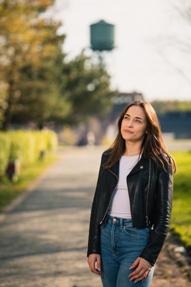 Frau in Lederjacke steht an einem Weg, im Hintergrund ein Wasserturm und Bäume.