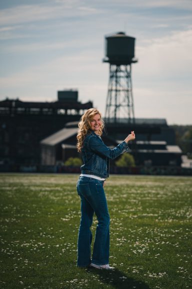 Frau mit lockigem Haar in Jeans auf einer Wiese, im Hintergrund ein Wasserturm.