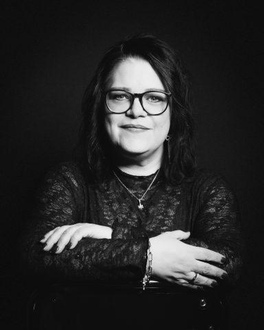 Porträt einer Frau mit Brille, die lächelt und die Arme gekreuzt hält.