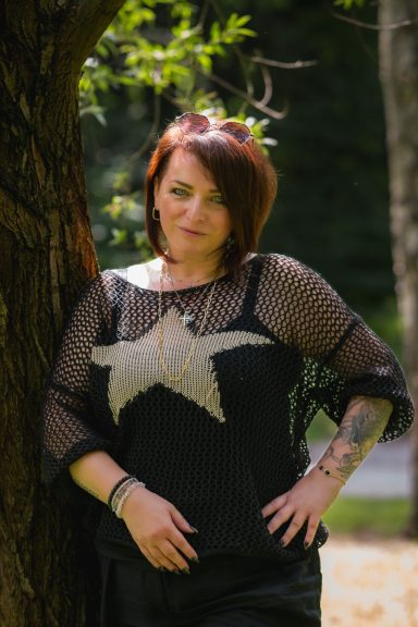 Frau mit kurzem, rotem Haar in einem Netzoberteil mit Sternmotiven, an einem Baum stehend.