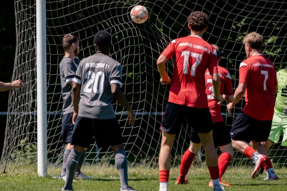 Fußballspieler in roten und grauen Trikots während eines Spiels, Ball fliegt ins Netz.