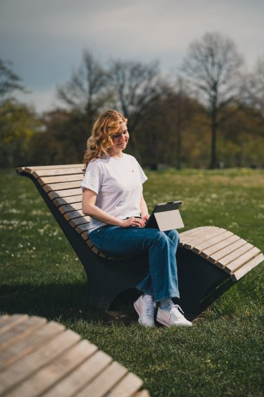 Eine Frau mit lockigen Haaren sitzt entspannt auf einer Bank im Park und hält ein Tablet.