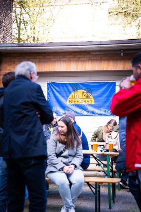 Personen stehen und sitzen bei einer Veranstaltung im Freien, mit Bier und einer Flagge im Hintergrund.