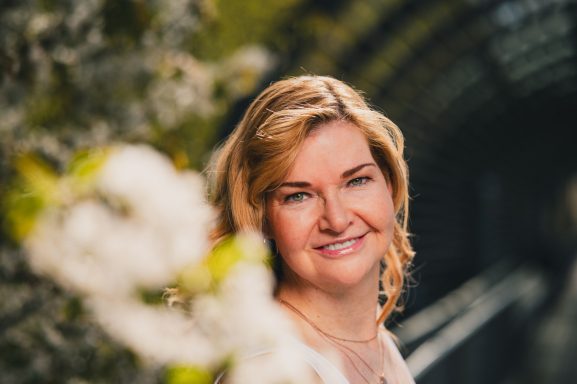 Lächelnde Frau mit blonden Haaren vor blühenden Pflanzen in einem alten Gebäude.