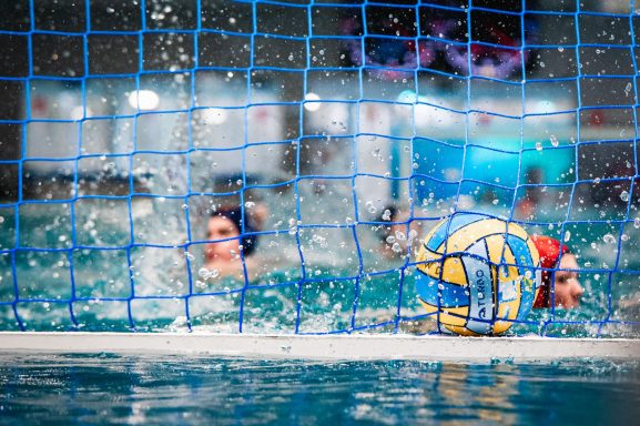 Wasserballspielerinnen im Sprung vor einem Tor, Wasser spritzt um sie herum.