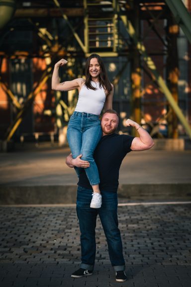 Ein Mann steht mit einer Frau auf seinen Schultern, beide zeigen muskulöse Arme.