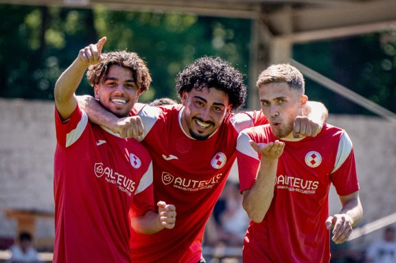 Drei Fußballspieler in roten Trikots posieren fröhlich und zeigen verschiedene Gesten.