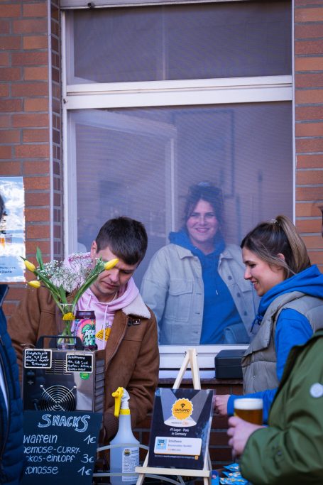 Ein Verkaufsstand mit zwei Personen, eine blickt durch ein Fenster, Blumen auf dem Tisch.