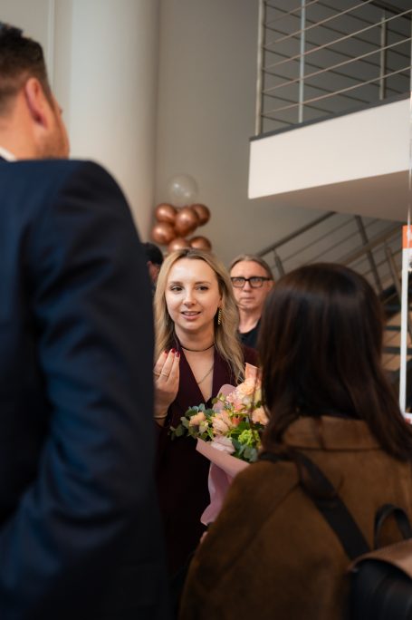 Frau mit Blumen in der Hand, umgeben von Personen in einem modernen Raum.
