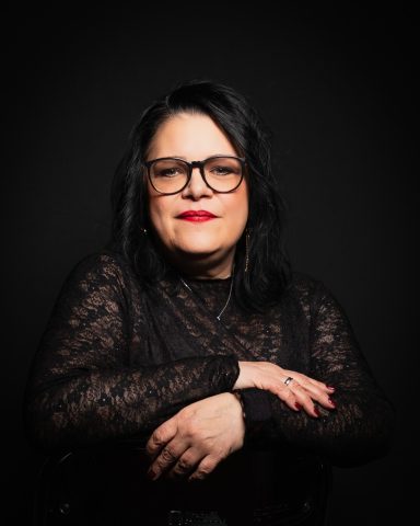 Frau mit dunklen Haaren und Brille, in schwarzer Spitze, mit verschränkten Armen.